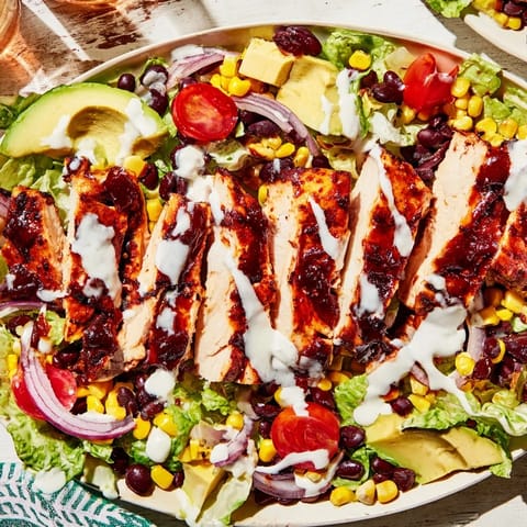 A hearty BBQ Chicken Salad features smoky grilled chicken, juicy cherry tomatoes, and ripe avocado slices, tossed with shredded cheese and a zesty lime ranch drizzle.
