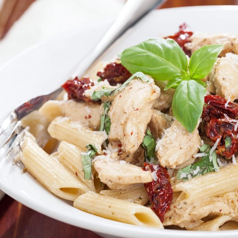 A close-up of Creamy Sun-Dried Tomato Chicken Pasta, highlighting glossy sauce clinging to penne with chicken and sun-dried tomatoes, garnished with fresh basil.  